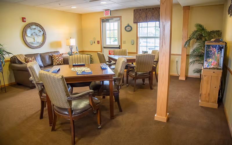 A cozy common area in a senior living facility with two wooden tables surrounded by cushioned chairs. There is a brown leather sofa with patterned pillows against the wall, a decorative wall hanging above it, a lamp on a side table, and a fish tank on a wooden stand near a window. The room has beige walls, carpeted floor, and a door with an exit sign above it.
