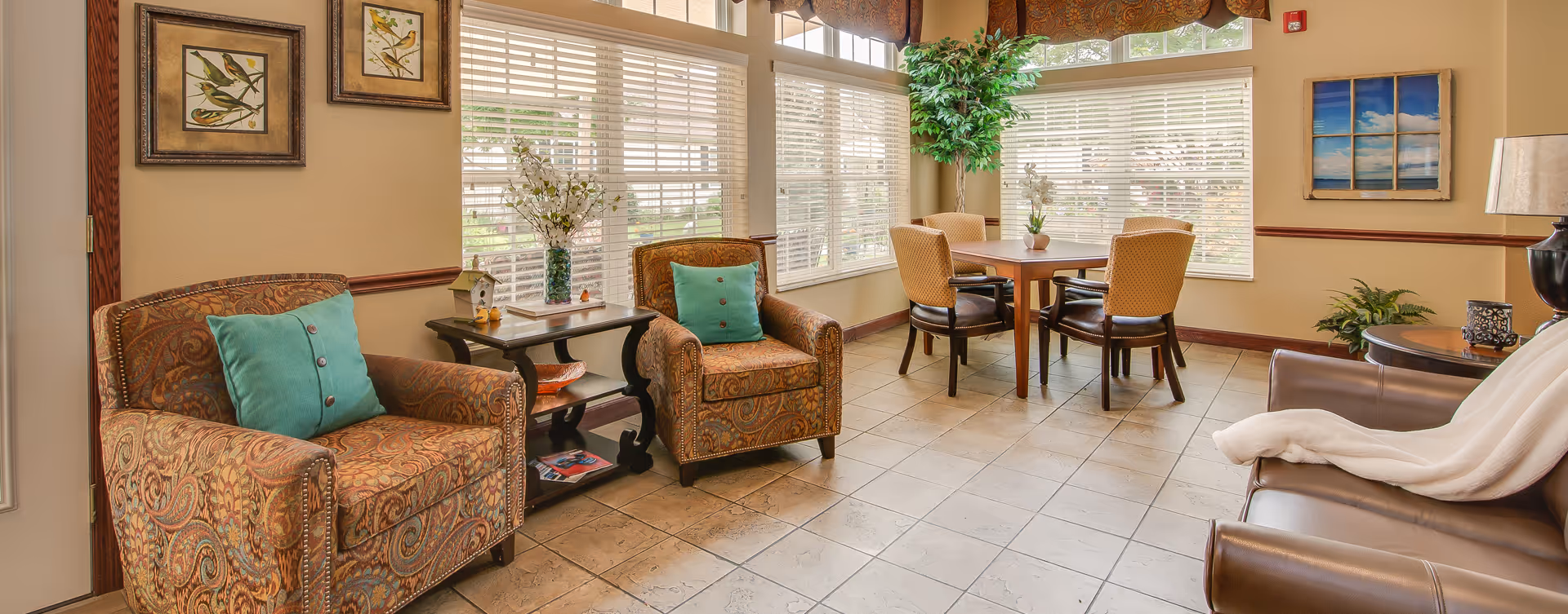 A cozy living room area with two patterned armchairs with teal cushions, a small dark wooden side table with decorative items, a square wooden table with four upholstered chairs near large windows with white blinds, a tall green plant in the corner, and a brown leather armchair with a white throw blanket. The walls are beige with framed artwork and a table lamp on a round side table.