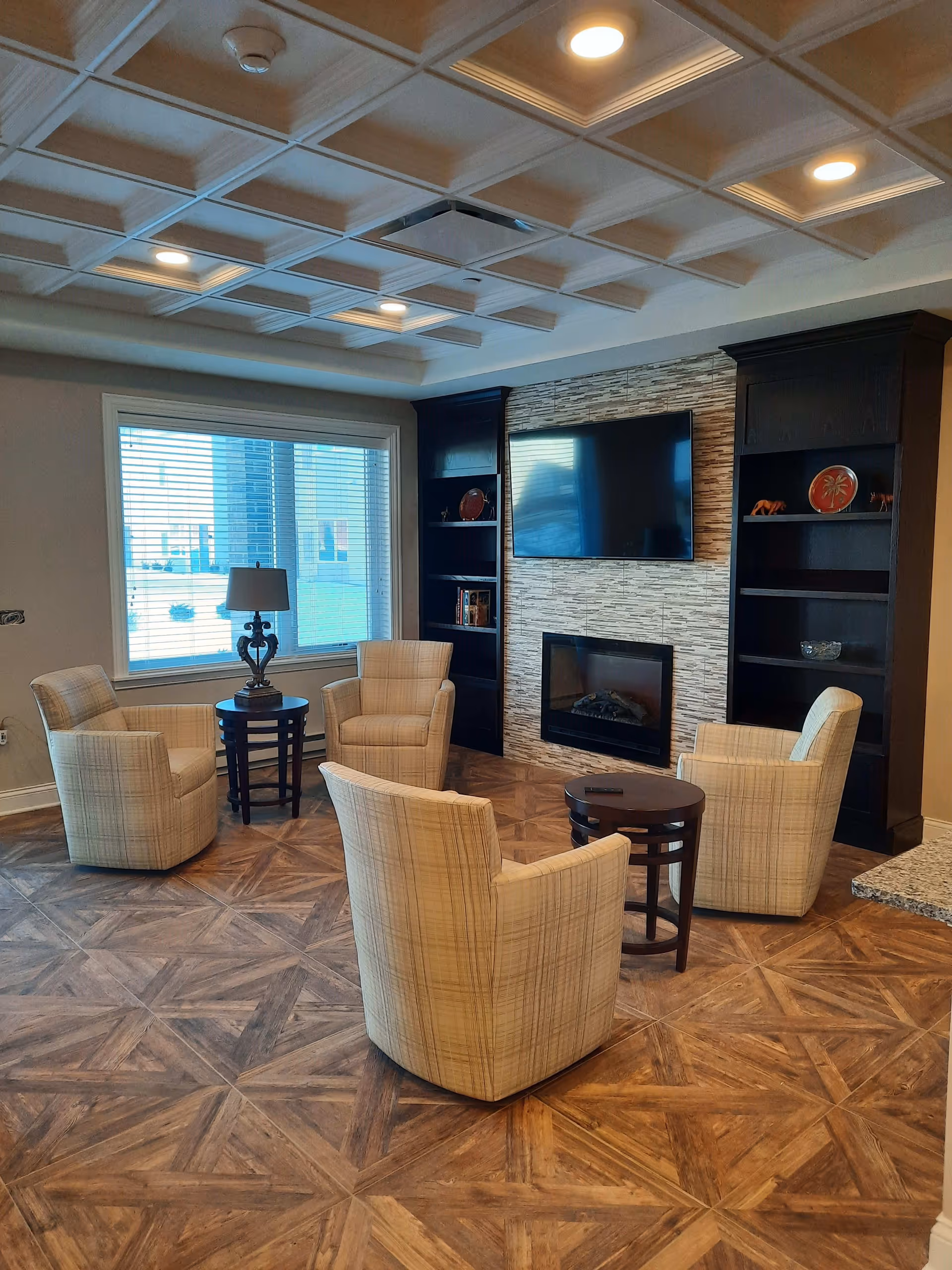 A cozy living room area with four beige plaid armchairs arranged around two small dark wooden tables. The room features a large window with blinds, a decorative coffered ceiling with recessed lighting, a wall-mounted flat-screen TV above a modern electric fireplace, and built-in dark wood shelves on either side of the fireplace.
