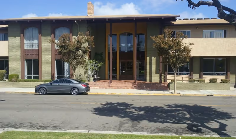 Front exterior view of a two-story building with large windows and a central entrance with tall glass doors. There are two small trees and a parked gray car in front of the building under a clear blue sky.