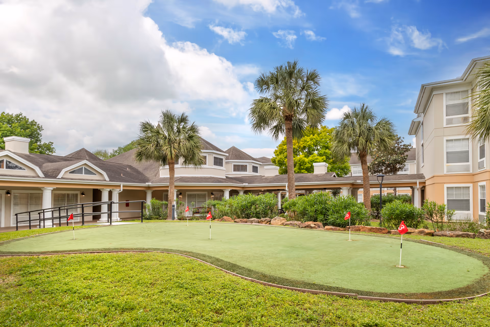 Outdoor putting green with several small red flags surrounded by grass and palm trees, with a multi-story senior living facility building in the background under a partly cloudy sky.