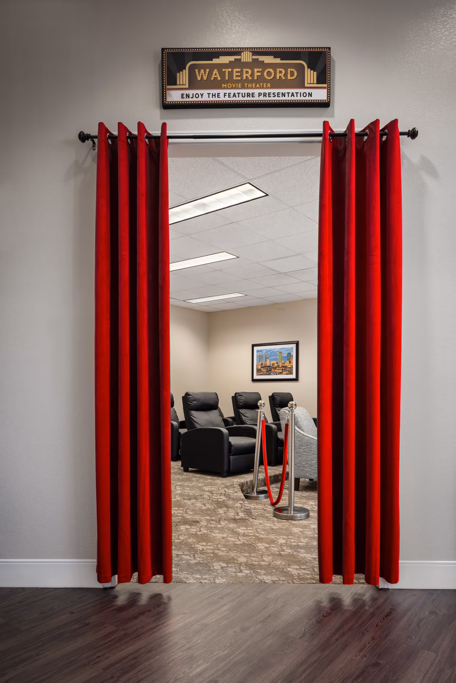 Entrance to a small movie theater room with red velvet curtains pulled to the sides. Inside, there are several black reclining chairs and a gray armchair arranged on a carpeted floor. A framed picture hangs on the beige wall, and a sign above the entrance reads 'Waterford Movie Theater Enjoy the Feature Presentation.'