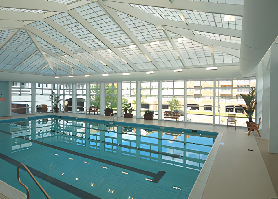 Indoor swimming pool with clear blue water, surrounded by large windows and a glass ceiling allowing natural light to fill the space. There are a few potted plants and chairs along the poolside.