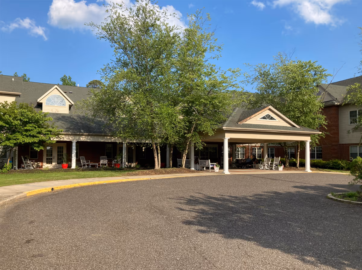 Exterior view of Woodview Estates facility showing a covered entrance with white columns, surrounded by trees and greenery under a blue sky with some clouds. The building has a brick and siding facade with multiple windows and a paved driveway in front.