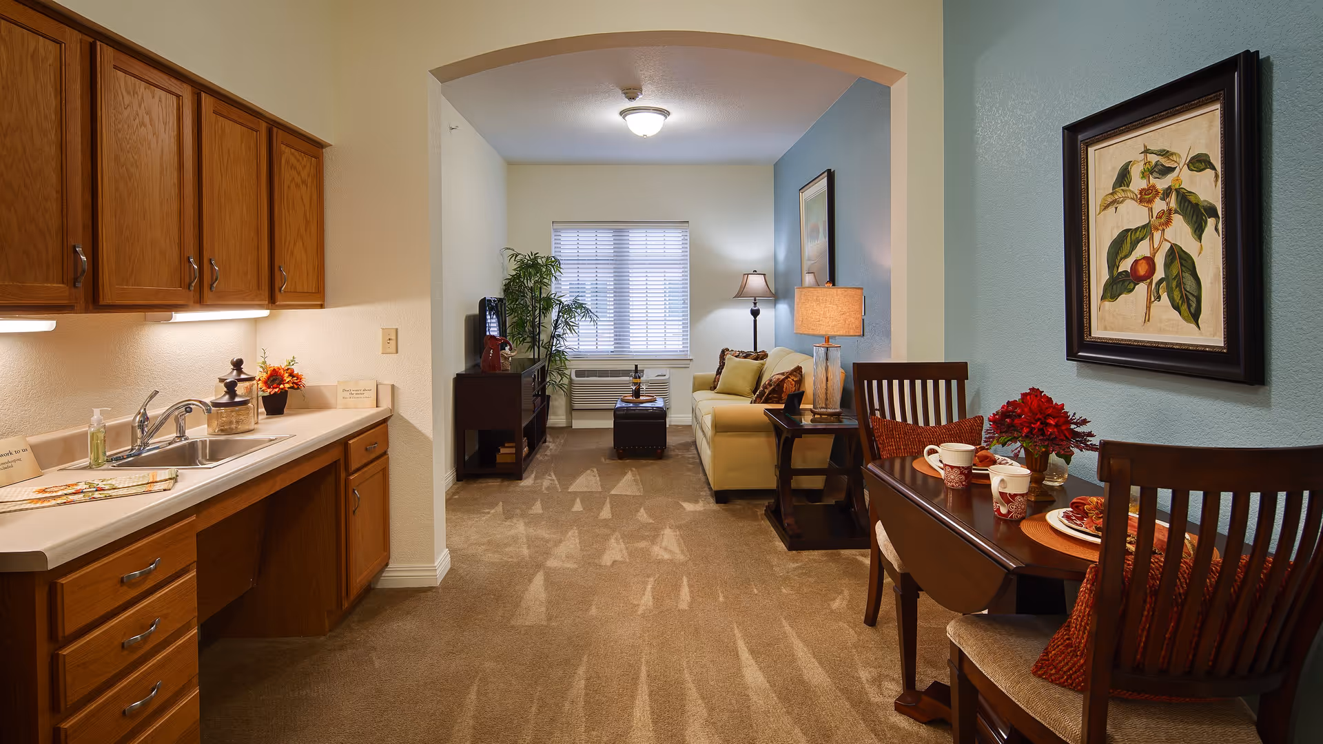 Interior view of a senior living apartment featuring a small kitchen area with wooden cabinets and a sink on the left, a dining table with two chairs and place settings on the right, and a living room area with a sofa, side table with a lamp, TV stand, and a window with blinds in the background. The walls are painted in light colors with a framed botanical artwork above the dining table.