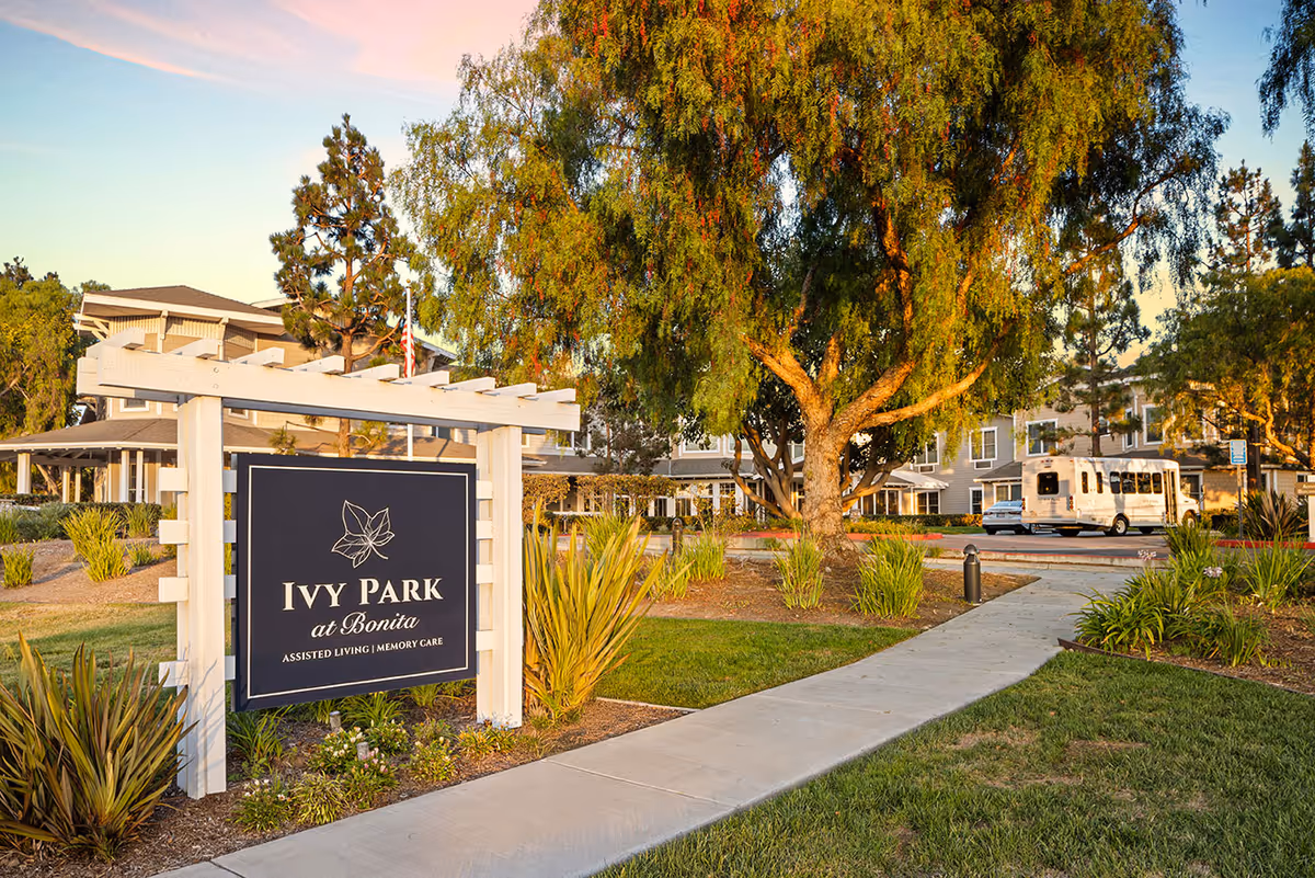 Outdoor view of Ivy Park at Bonita senior living facility sign with landscaped garden, trees, a sidewalk, and the building in the background during sunset.