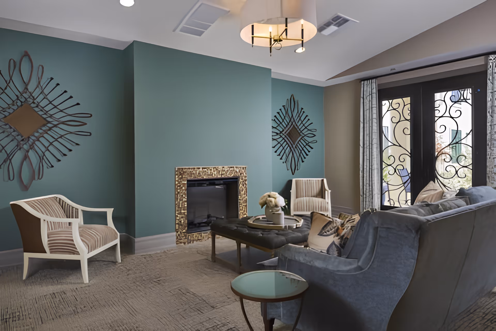 A cozy living room with teal and beige walls, featuring a fireplace with a decorative gold frame, two armchairs with striped upholstery, a gray sofa with patterned pillows, a tufted ottoman with a tray and a small decorative item, a round side table, and French doors with ornate wrought iron designs leading outside.