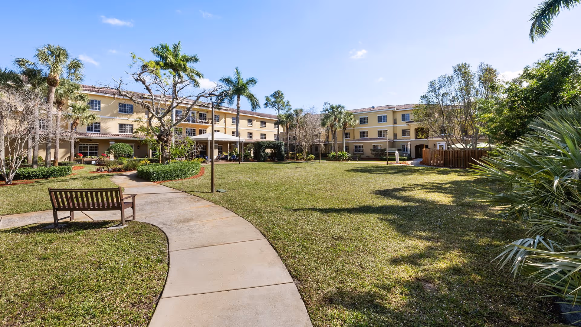 A spacious outdoor courtyard area at Atria Park of St. Josephs featuring a curved concrete walkway, a wooden bench, well-maintained grassy lawn, palm trees, and a three-story beige building in the background under a clear blue sky.