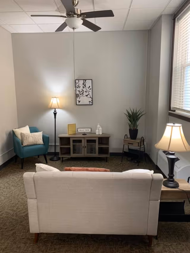 A cozy living room with a beige sofa facing a wooden cabinet against a light gray wall. To the left is a teal armchair with a pillow, and a floor lamp beside it. On the right side, there is a small table with a potted plant and another table lamp. A ceiling fan is visible above, and a window with blinds is on the right wall.
