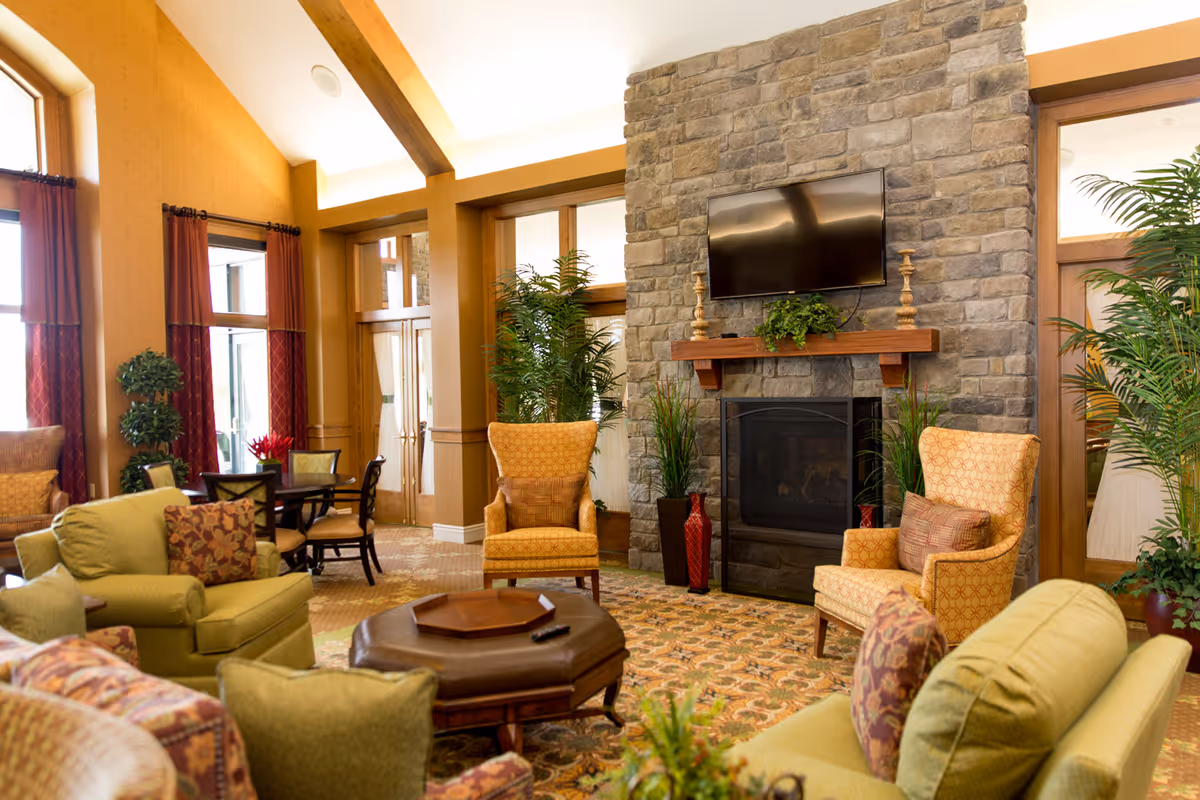 A cozy living room area in a retirement community featuring a stone fireplace with a mounted flat-screen TV above it. The room is furnished with comfortable armchairs and sofas in warm tones, a wooden coffee table, and decorative plants. Large windows with red curtains allow natural light to fill the space.