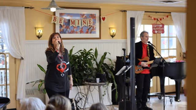 A woman singing into a microphone and a man playing an electric guitar and singing in a decorated room with a 'Be Mine' banner and Valentine's Day decorations. There are plants and a piano in the background, and an audience is partially visible in the foreground.