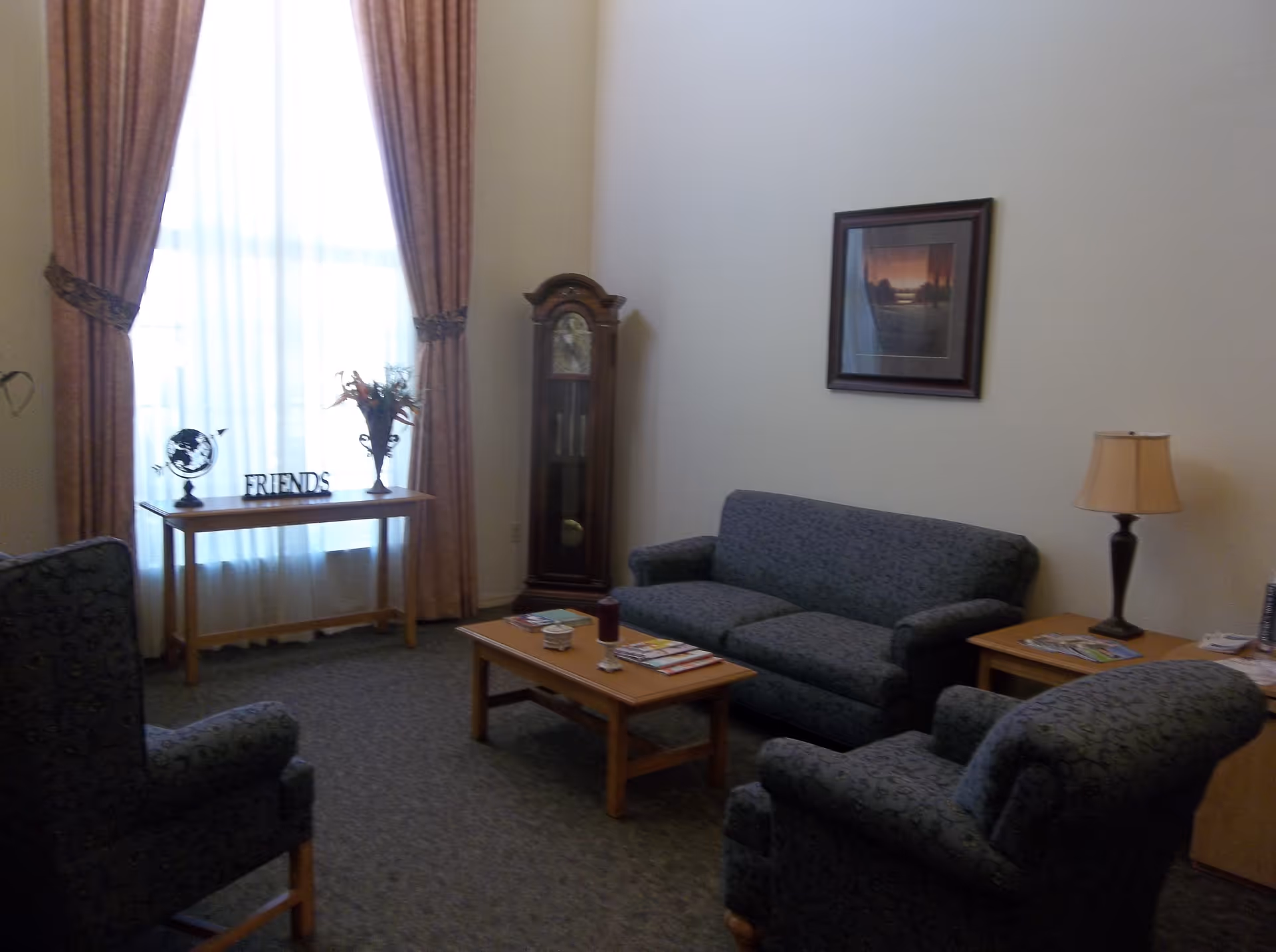 A cozy living room with a sofa and two armchairs upholstered in dark patterned fabric. A wooden coffee table with magazines and a candle sits in the center. A wooden side table with a lamp and more magazines is next to the sofa. A tall grandfather clock stands against the wall near a large window with sheer white curtains and heavy drapes. A wooden console table under the window holds a globe, a vase with flowers, and a decorative sign that says 'FRIENDS'. A framed picture hangs on the wall above the sofa.