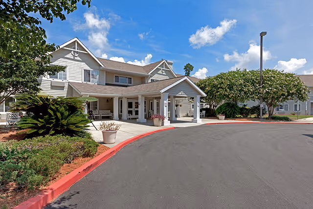 Exterior view of a senior living facility building with a covered entrance, surrounded by landscaped greenery and a curved driveway under a partly cloudy blue sky.