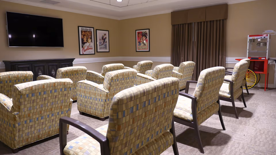 A small theater-style room with two rows of cushioned chairs facing a large flat-screen TV mounted on the wall. The walls are beige with white trim, decorated with framed classic movie posters. There is a popcorn machine on a red cart in the corner near a window with brown curtains.
