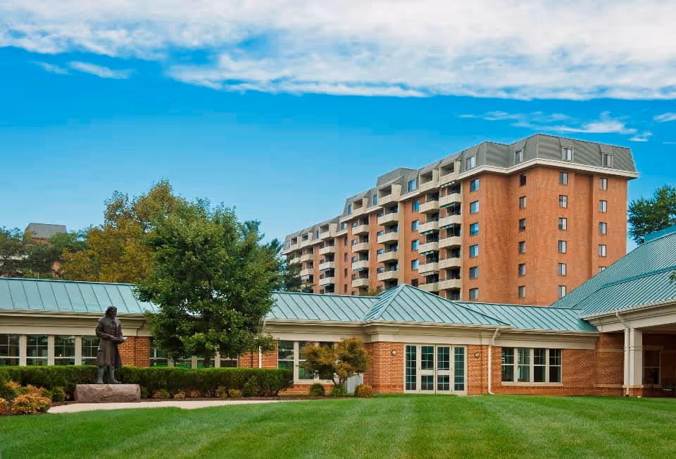 Exterior view of Asbury Solomons Island facility showing a multi-story brick building with balconies, a single-story building with green metal roofing, a well-maintained lawn, trees, and a statue on a stone pedestal.