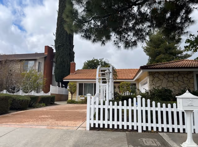 Front exterior of a single-story house with a white picket fence, brick driveway, arched trellis, and surrounding trees.