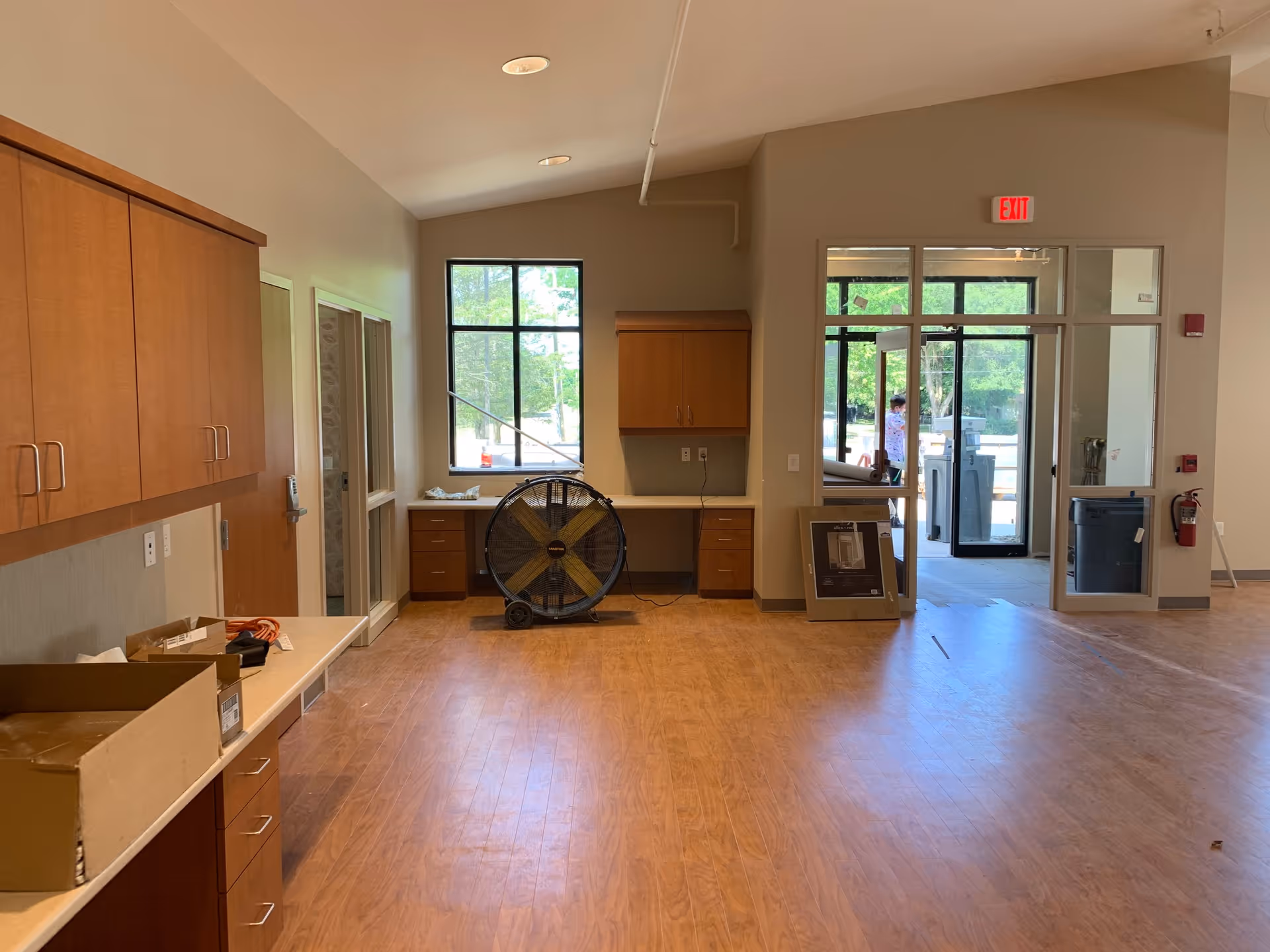 Interior view of a room with wooden cabinets and drawers along the left and back walls, a large industrial fan on the floor near a window, and a glass door leading outside. The floor is wooden, and there are some boxes and items on the counter. The room appears to be under setup or renovation.