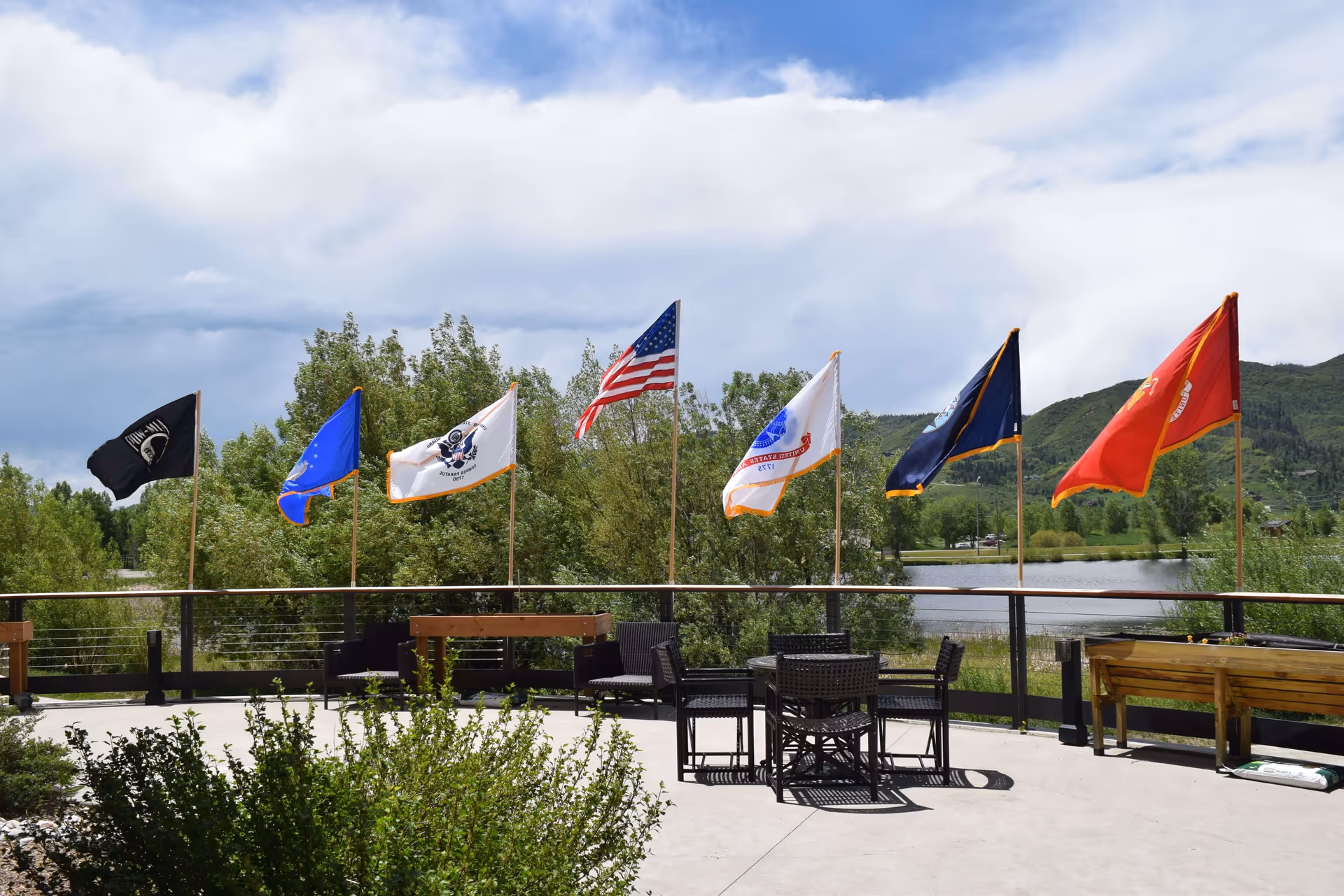 Outdoor patio with tables and chairs and a row of flags flying beside a lake and trees.