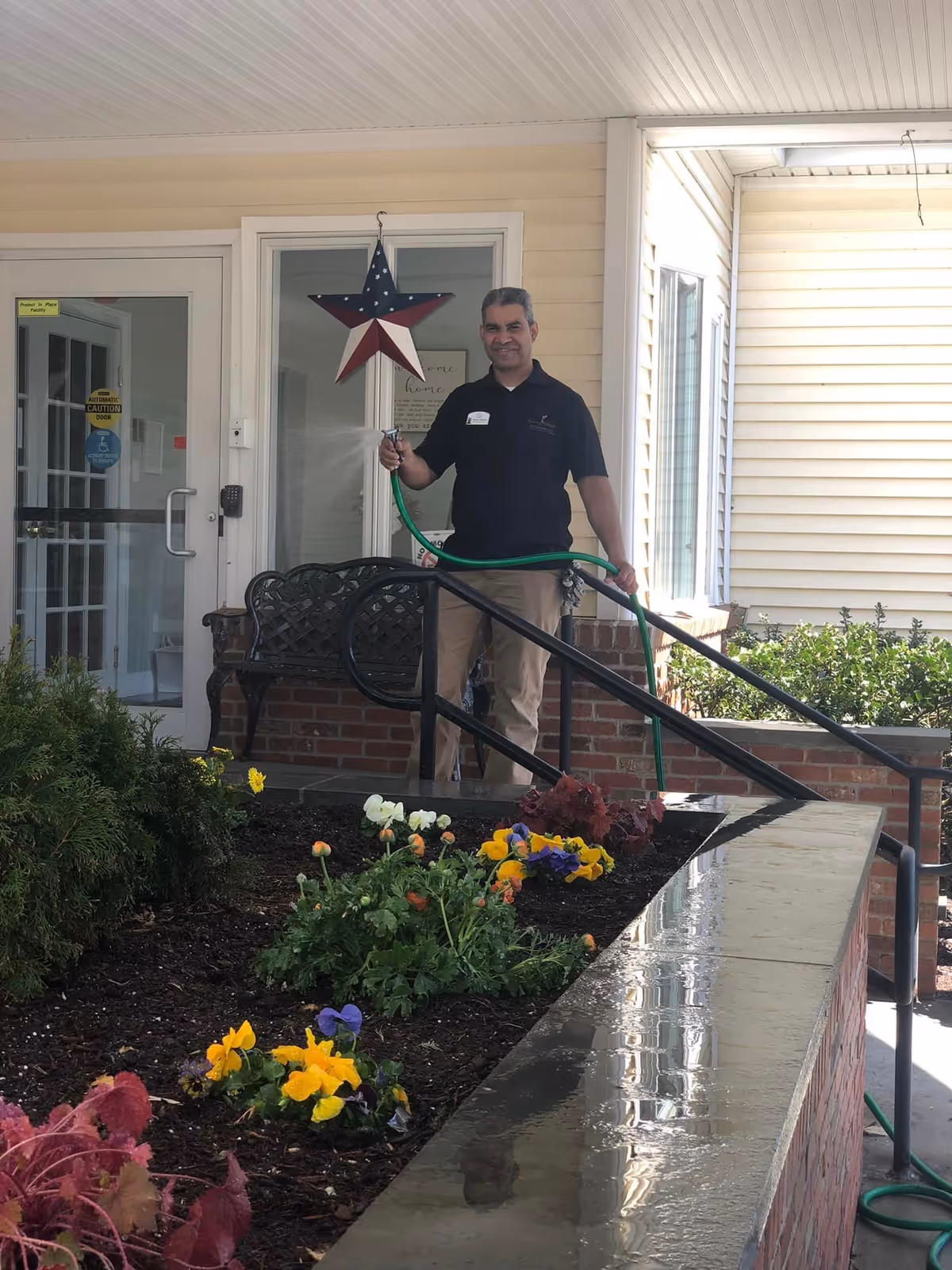 A man standing outside a building holding a green garden hose, watering a flower bed with colorful flowers. The building has beige siding, a glass door, and a decorative star hanging on the wall. There is a black metal bench and a brick planter with flowers in front of the man.