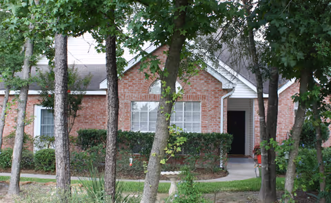 Front exterior view of a single-story brick cottage with white trim, partially obscured by several trees and shrubs. The cottage has a gabled roof, a window with white blinds, and a black front door with a small covered porch.