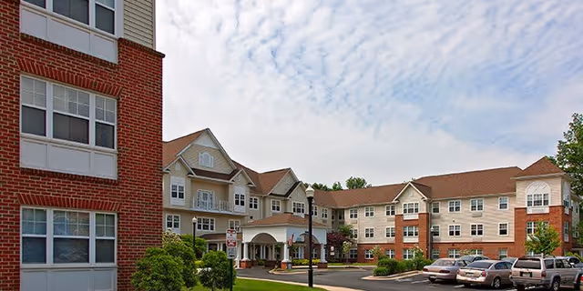 Exterior view of Hampshire Village senior living facility showing a multi-story building with a combination of brick and beige siding, a covered entrance, several windows, a parking lot with cars, and a partly cloudy sky.