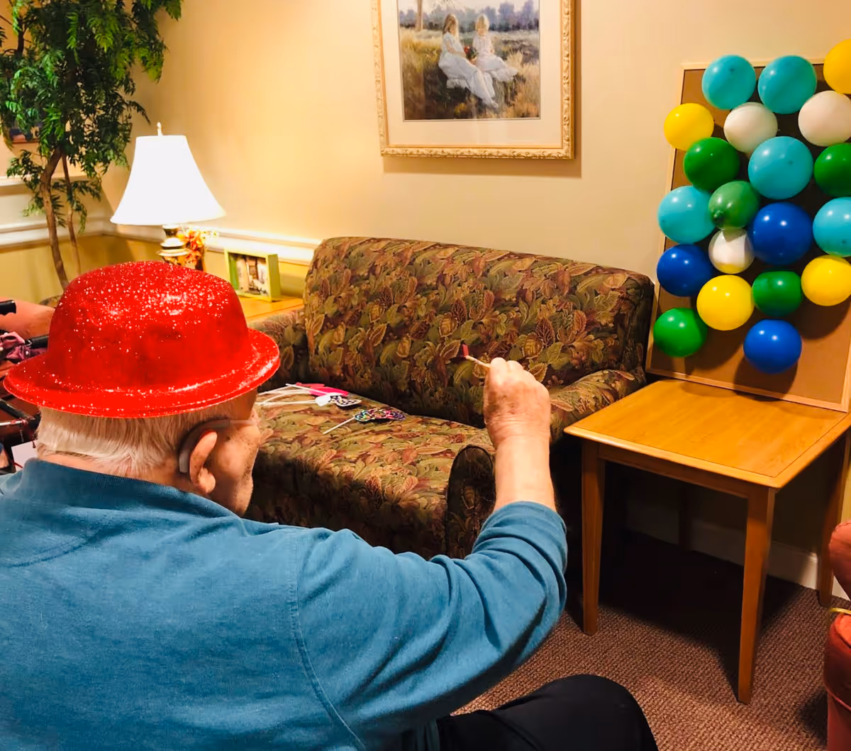 An elderly person wearing a red glittery hat is playing a balloon dart game indoors. The person is aiming a dart at a board with colorful balloons attached, set on a wooden table. The room has a floral-patterned couch, a lamp, a framed picture on the wall, and a potted plant in the corner.