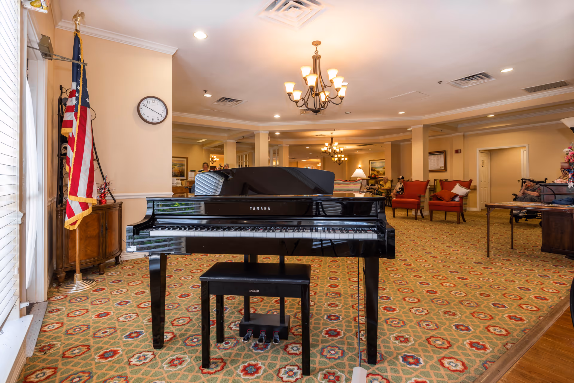 Interior view of a senior living facility lounge area featuring a black Yamaha grand piano with a matching bench in the foreground. The room has patterned carpet, beige walls, and multiple chandeliers hanging from the ceiling. There are several red armchairs and a few elderly residents seated in the background. An American flag stands near a window on the left side, and a clock is mounted on the wall above a wooden cabinet.