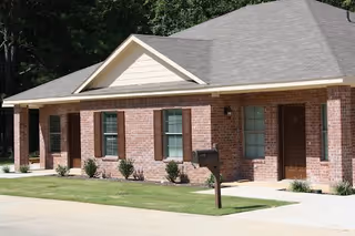 Exterior view of a single-story brick building with a gray shingled roof, several windows with brown shutters, a brown door, a mailbox on a post, and a concrete walkway surrounded by grass and small shrubs.