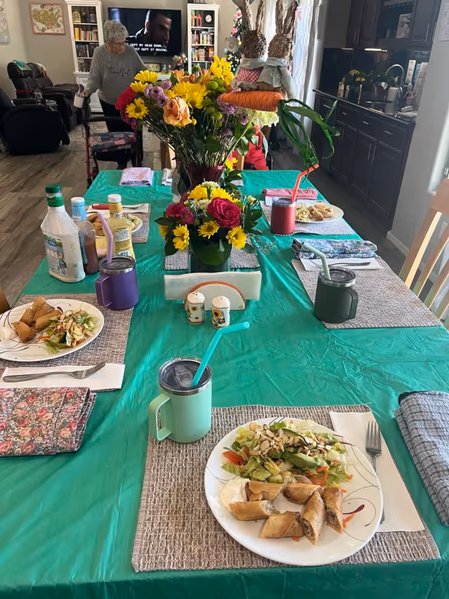 A dining table set for a meal with plates of salad and spring rolls, colorful flowers in vases as centerpieces, various condiments, and insulated mugs with straws. In the background, an elderly woman with a walker is visible near a living room area with a TV and bookshelves.