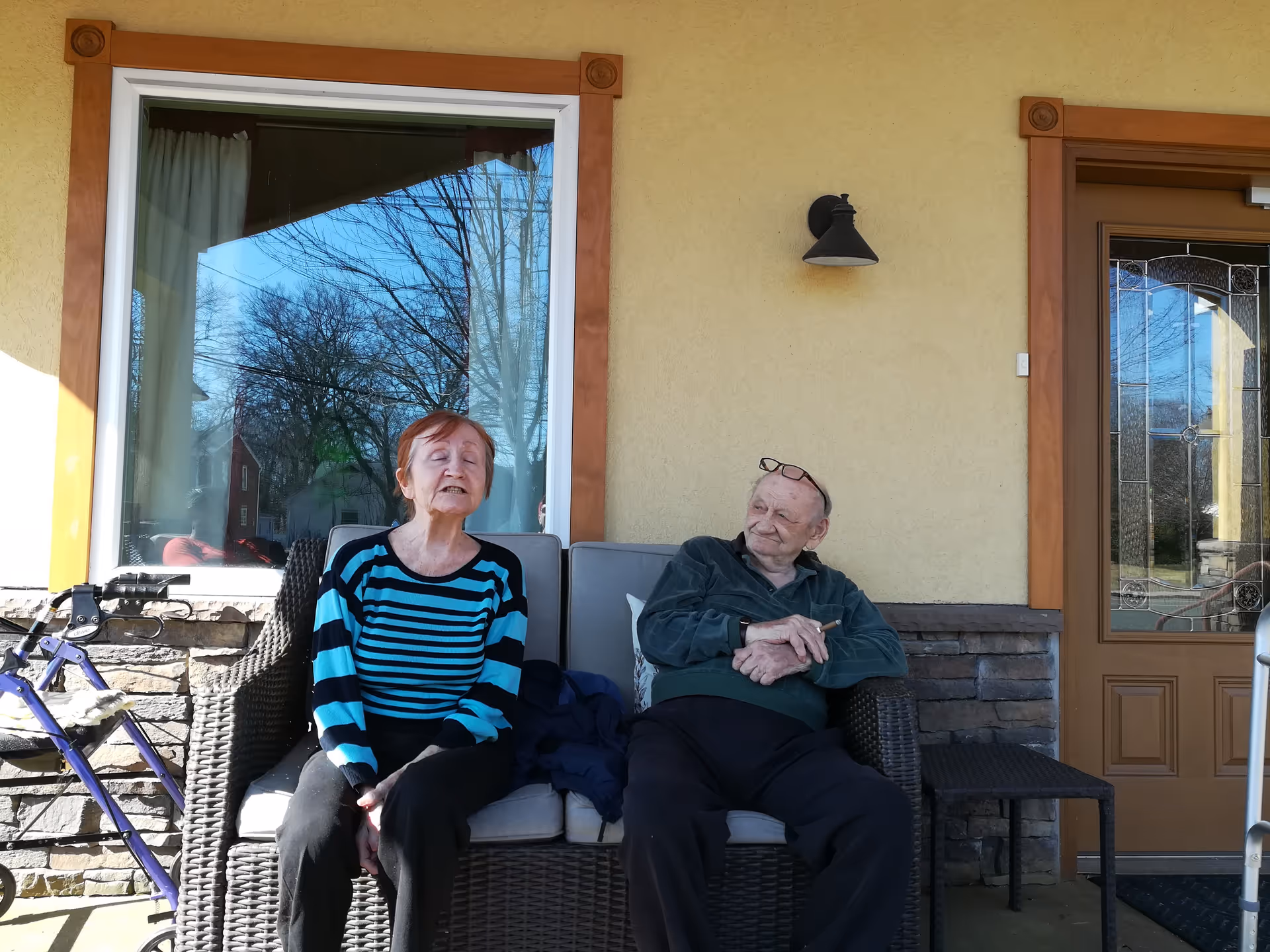 Two elderly individuals sitting on a wicker outdoor sofa on a porch. One person is wearing a blue and black striped sweater and black pants, and the other is wearing a dark jacket and black pants with glasses resting on their head. Behind them is a window with reflections of trees and a door with decorative glass panels. A walker is visible to the left of the sofa.