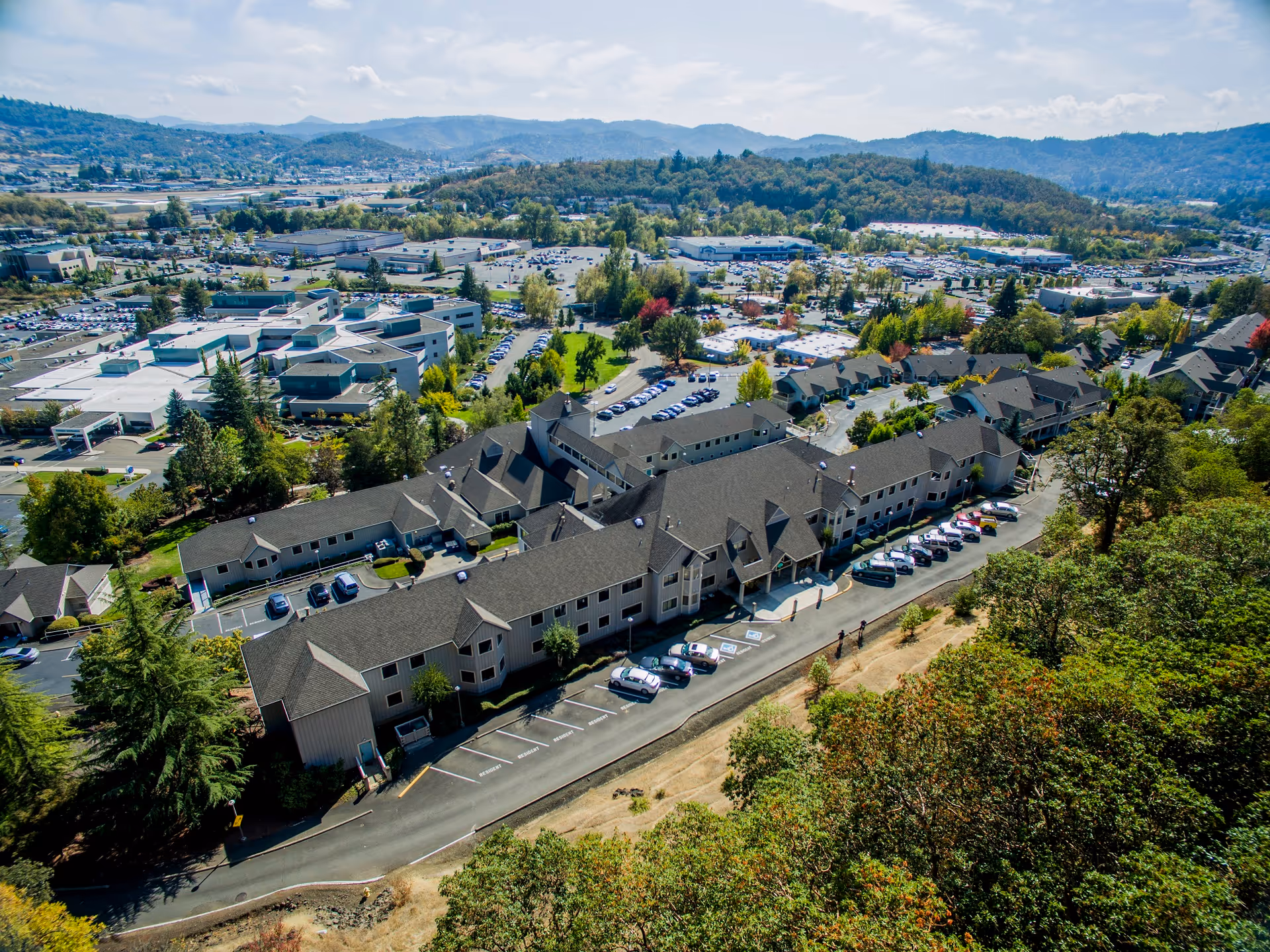 Aerial view of Linus Oakes Village, showing a large senior living facility with multiple connected buildings surrounded by trees and parking lots. The facility is situated in a suburban area with hills and other buildings in the background under a partly cloudy sky.