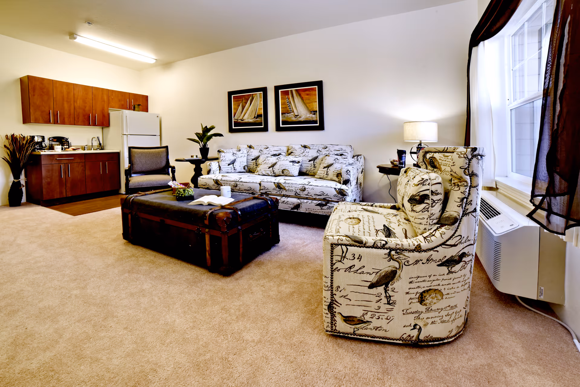 A cozy living room area in an assisted living facility featuring a patterned armchair and matching sofa with bird and script designs, a dark wooden trunk used as a coffee table, a side table with a lamp, and a small kitchenette with wooden cabinets, a refrigerator, and a sink in the background.