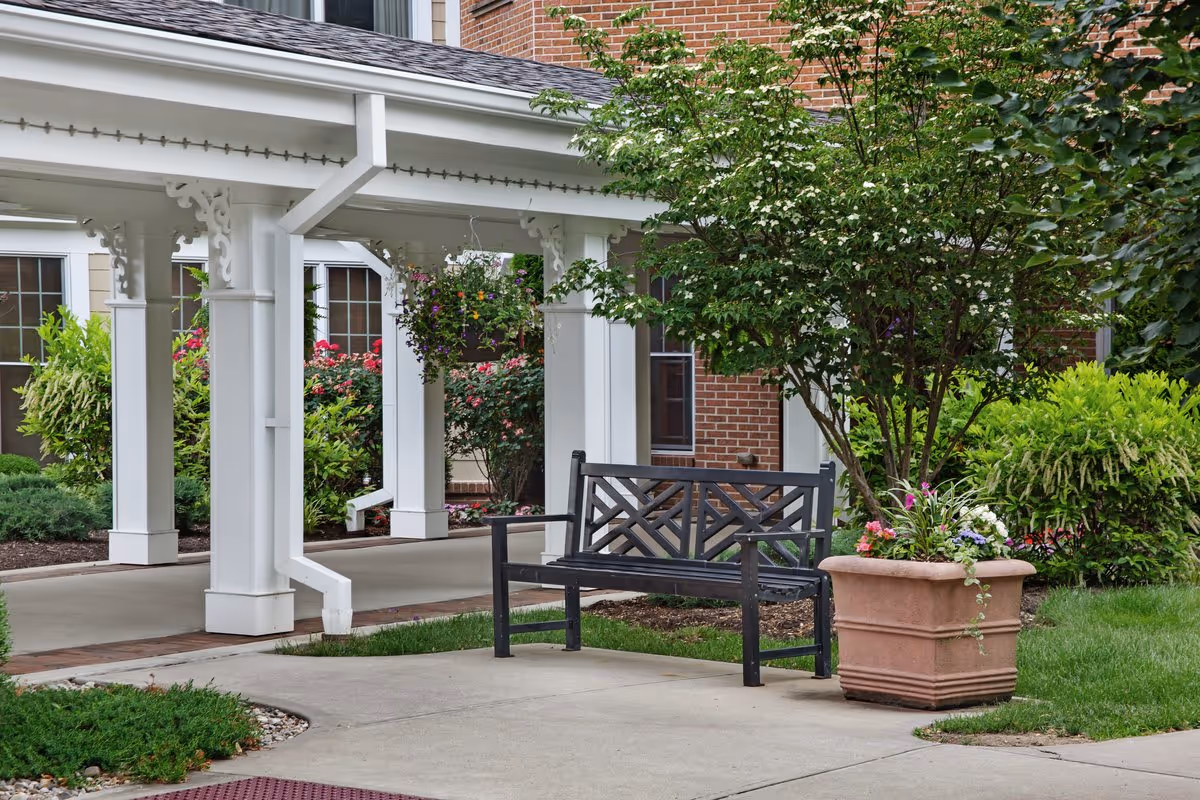 Outdoor seating area at Sunrise of Louisville featuring a black metal bench next to a large planter with colorful flowers and a small tree. The area is surrounded by green shrubs and plants, with a covered walkway and brick building in the background.