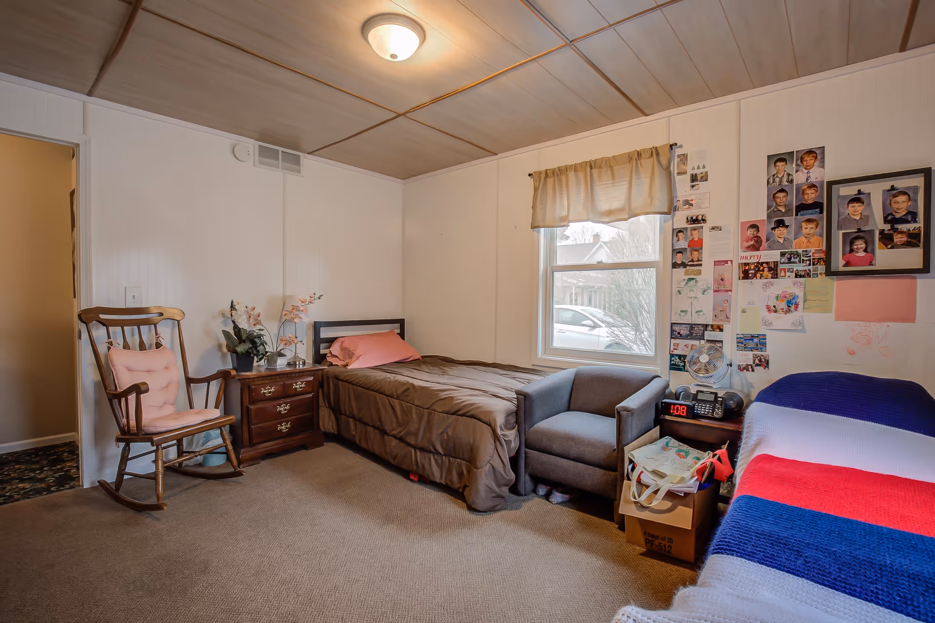 A cozy bedroom with two beds, one with a brown comforter and a pink pillow, and the other covered with a knitted blanket in blue, white, and red stripes. There is a wooden rocking chair with a pink cushion, a wooden nightstand with a lamp and flowers, a gray armchair, and a small table with a clock, radio, and fan. The wall next to the window is decorated with numerous photos and drawings. The room has beige carpet and a wooden paneled ceiling.