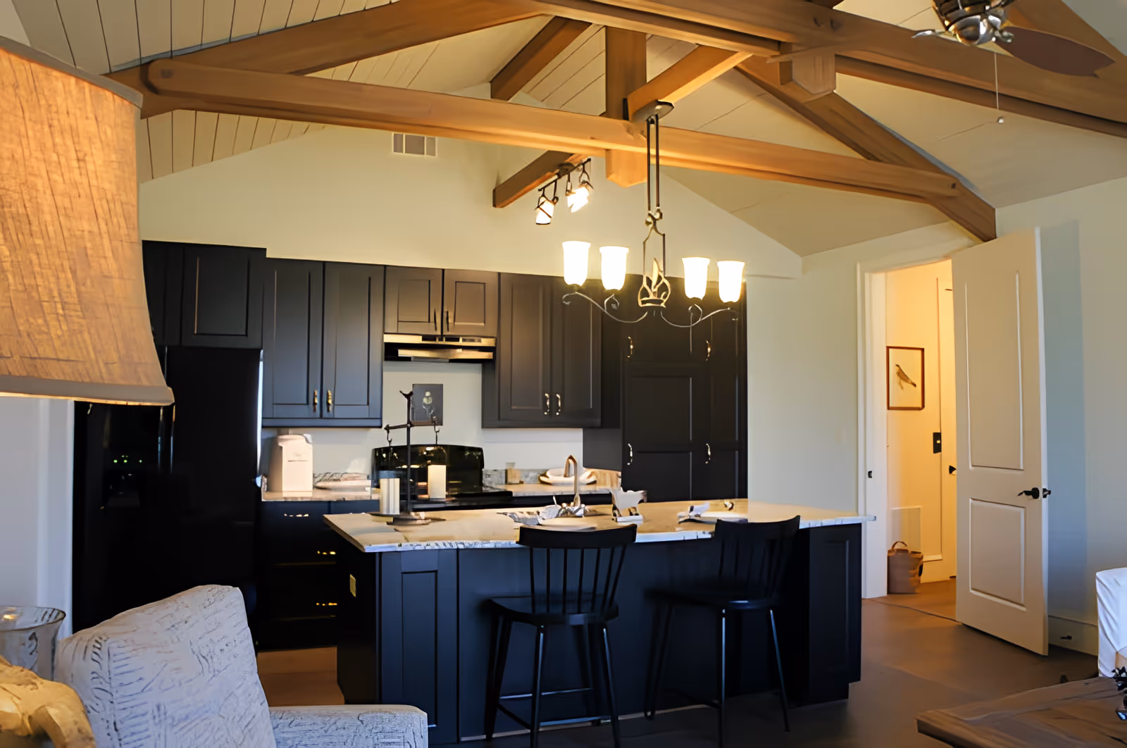 Interior view of a modern kitchen with dark cabinetry and a marble countertop island with two black bar stools. The ceiling features exposed wooden beams and a ceiling fan. A chandelier with multiple lights hangs above the island. To the right, an open door leads to another room with a framed picture on the wall.