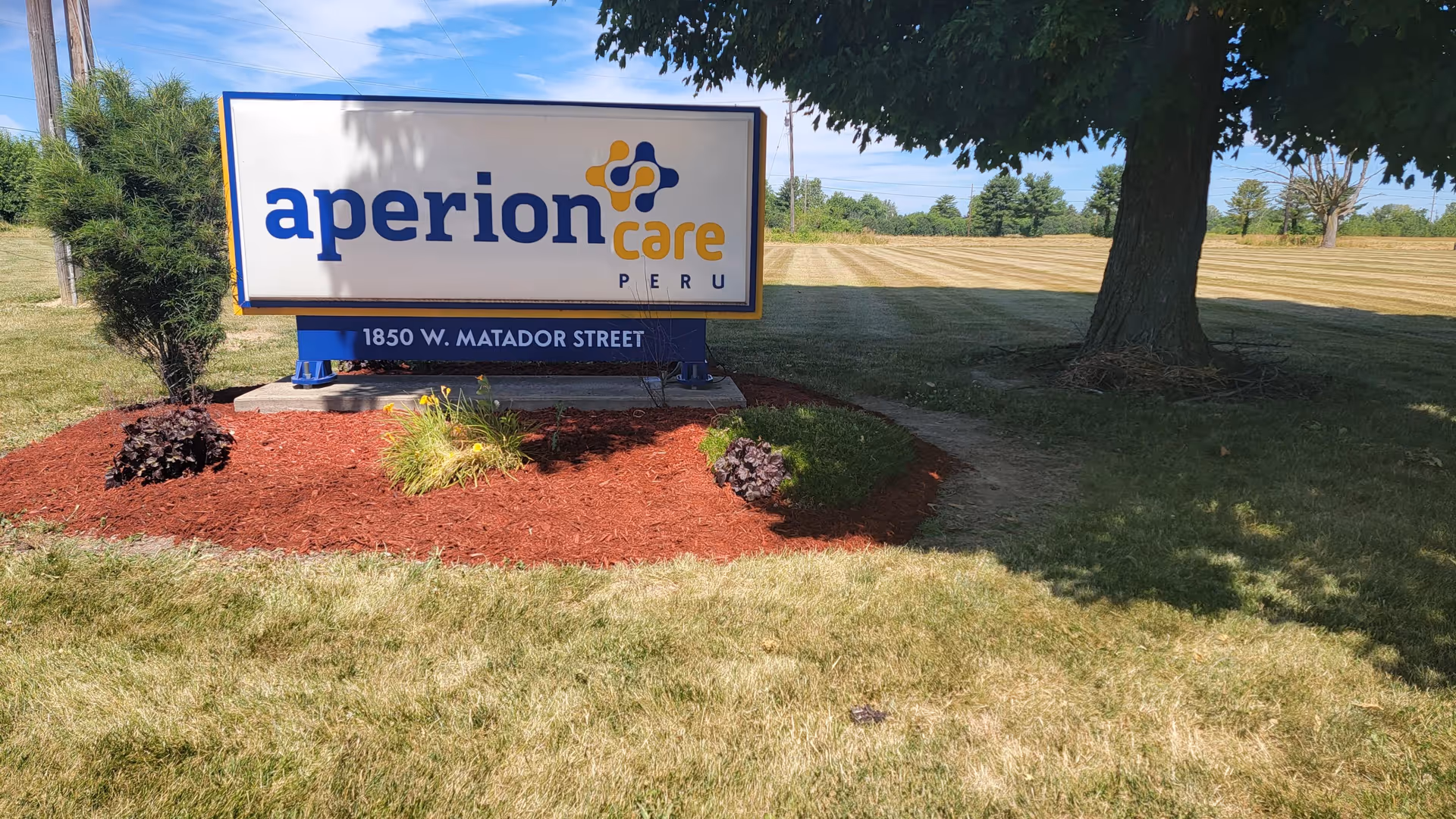 Outdoor view of a sign for Aperion Care Peru located at 1850 W. Matador Street, surrounded by grass, mulch, small plants, and a large tree providing shade.