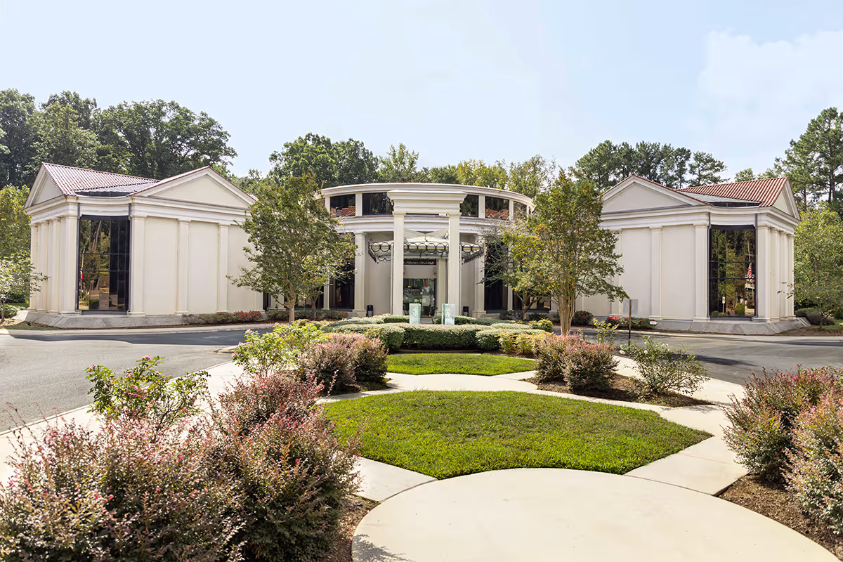 Front entrance of a classical-style building with columns, a circular drive and landscaped lawns and shrubs.