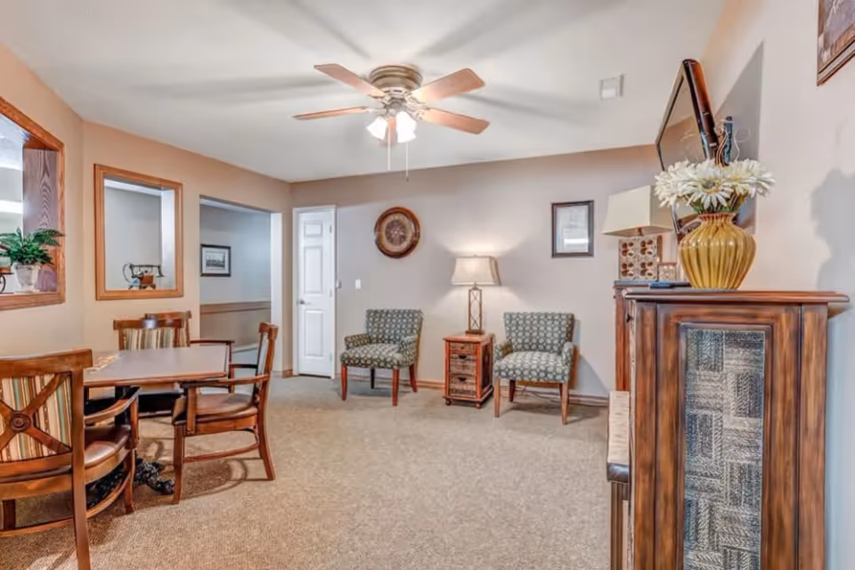 Comfortable communal living area with a dining table and chairs, two upholstered armchairs, a TV on a wooden cabinet, and a ceiling fan.