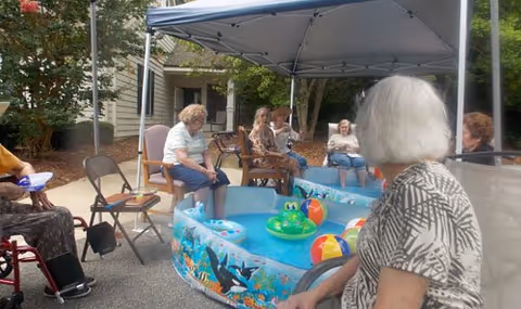 A group of elderly people sitting around a small inflatable pool filled with water and colorful beach balls under a canopy in an outdoor setting at an assisted living facility.