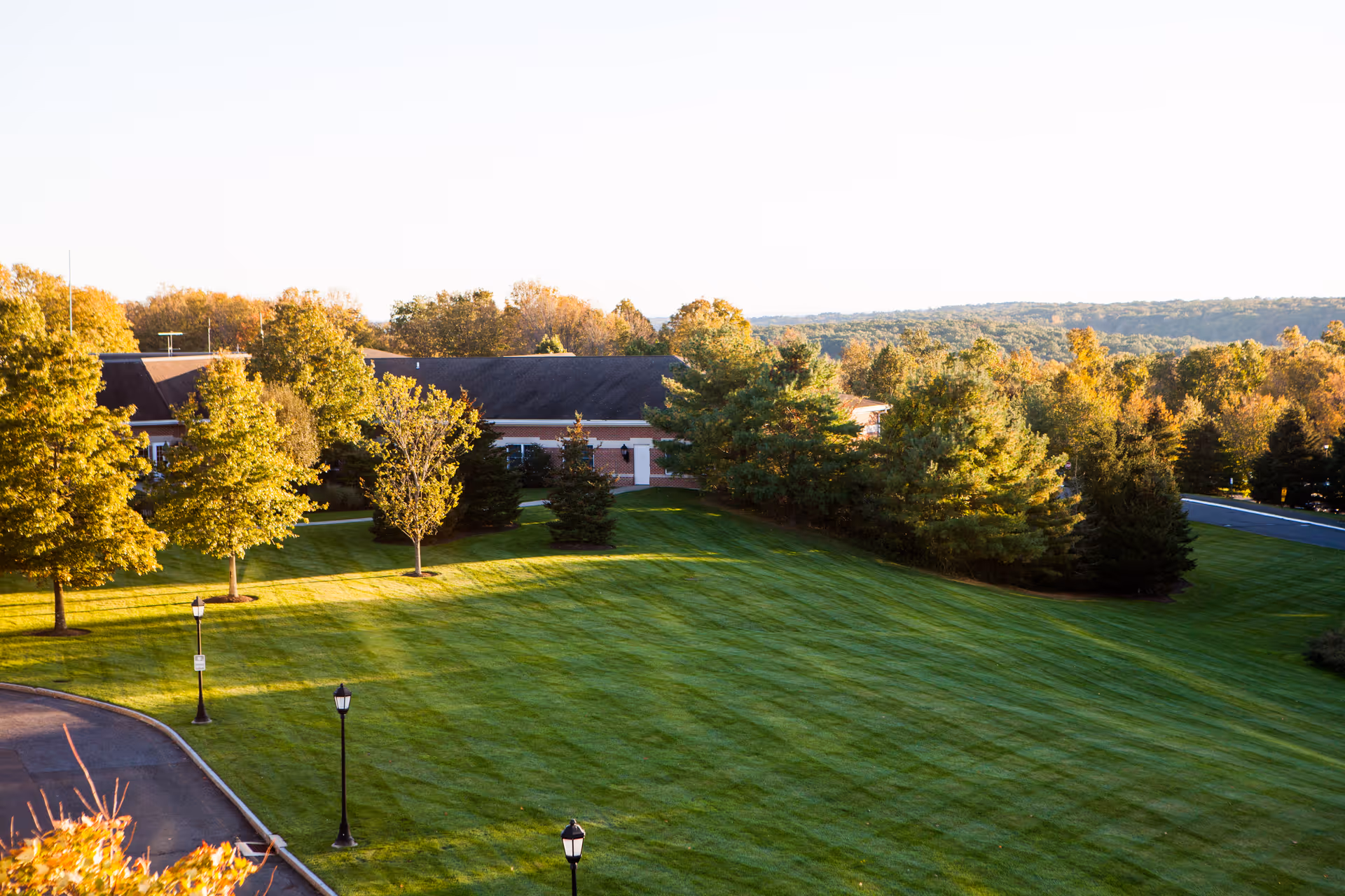 A large, well-maintained grassy lawn with several trees and lamp posts, adjacent to a single-story building with a dark roof. The background shows a scenic view of a forested area under a clear sky.