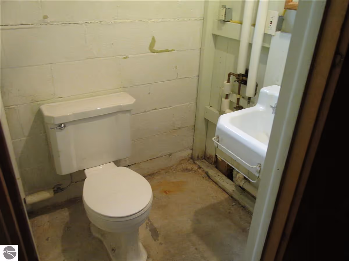 Small unfinished bathroom with a toilet, a wall-mounted sink, exposed plumbing, and a concrete floor with cinderblock walls.