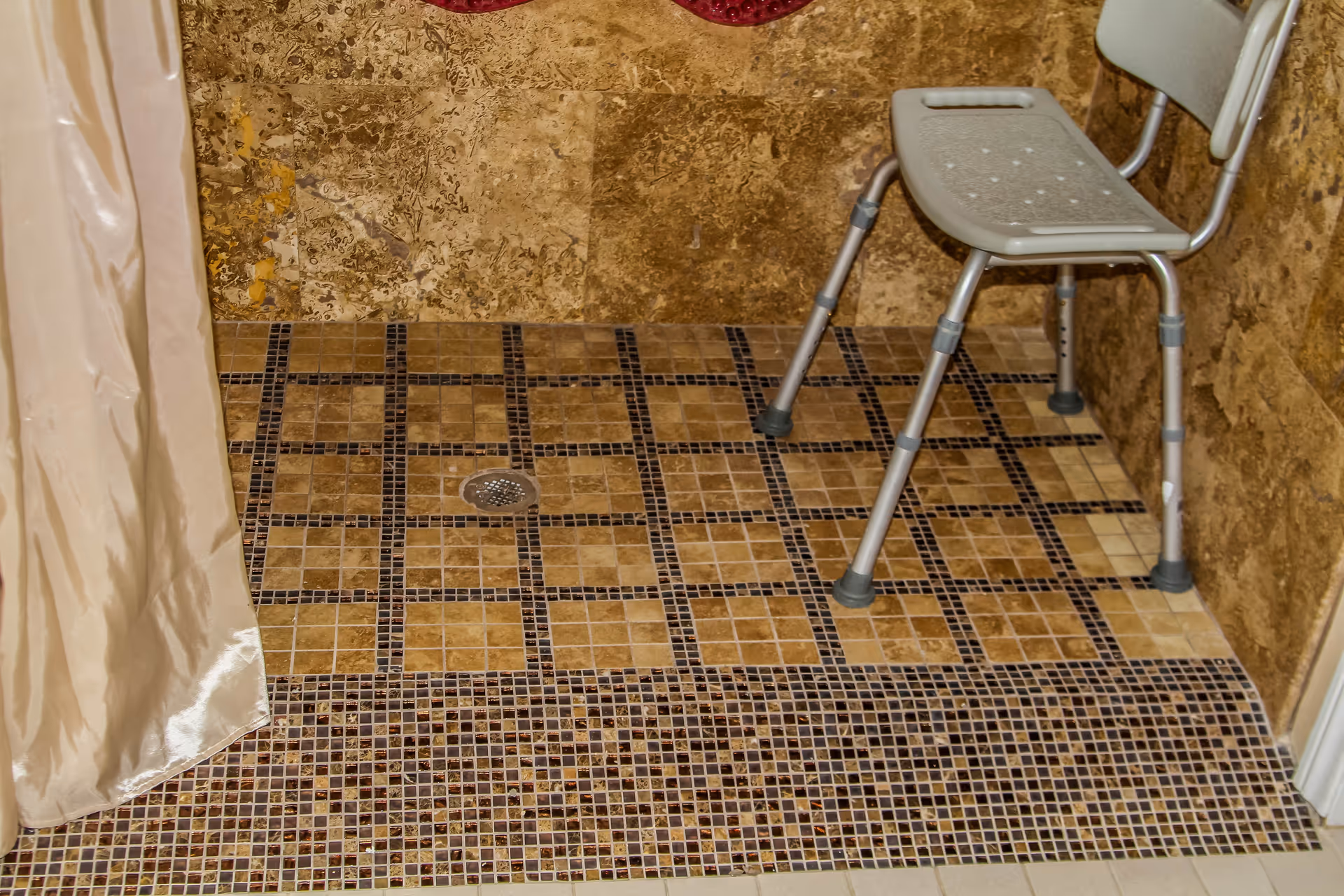 Shower area with brown and beige mosaic tile floor and walls, featuring a beige shower chair and a beige shower curtain partially visible on the left side.