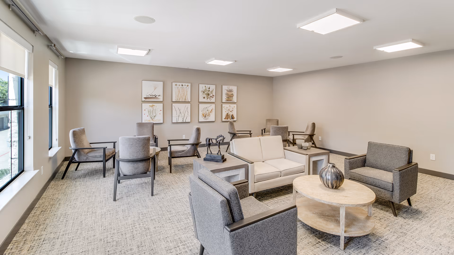A bright and spacious lounge area with multiple seating arrangements including armchairs and a loveseat around a wooden coffee table. The room has large windows with roller shades, neutral-colored walls adorned with framed botanical artwork, and modern ceiling lights.