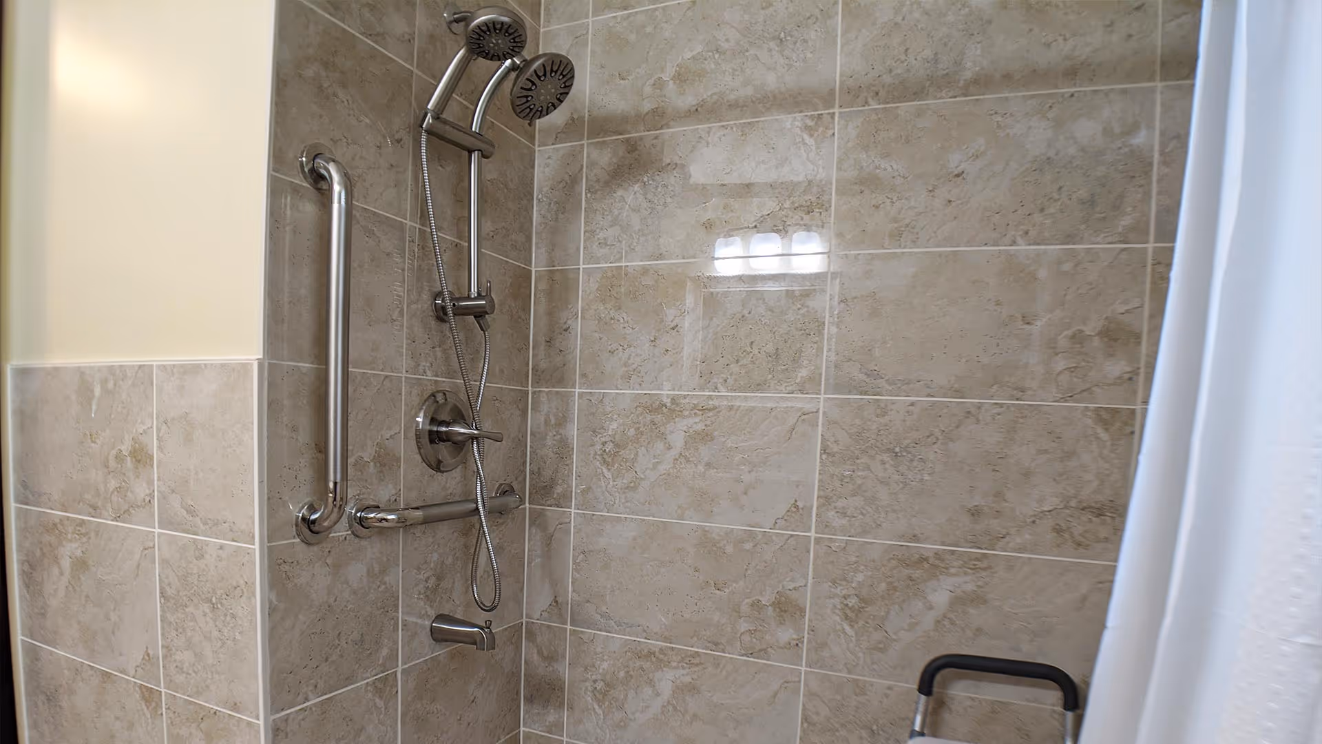 A tiled shower area with beige and light brown tiles, featuring a handheld showerhead attached to a vertical bar, a grab bar on the wall, and a white shower curtain partially visible on the right side.