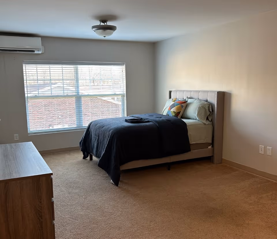 Bright bedroom with a bed against the right wall, a dresser on the left, and a large window letting in natural light.