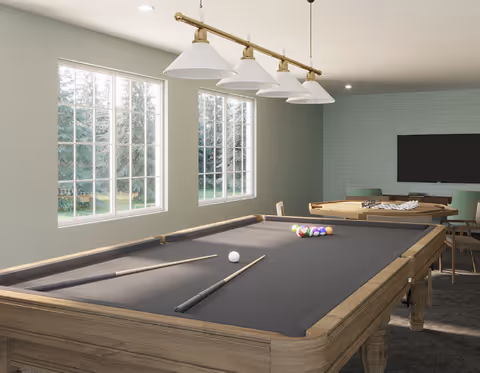 A well-lit game room with a pool table in the foreground, featuring pool cues and a rack of billiard balls. The room has large windows letting in natural light, a hanging light fixture above the pool table, and a TV mounted on the far wall with chairs and a table nearby.