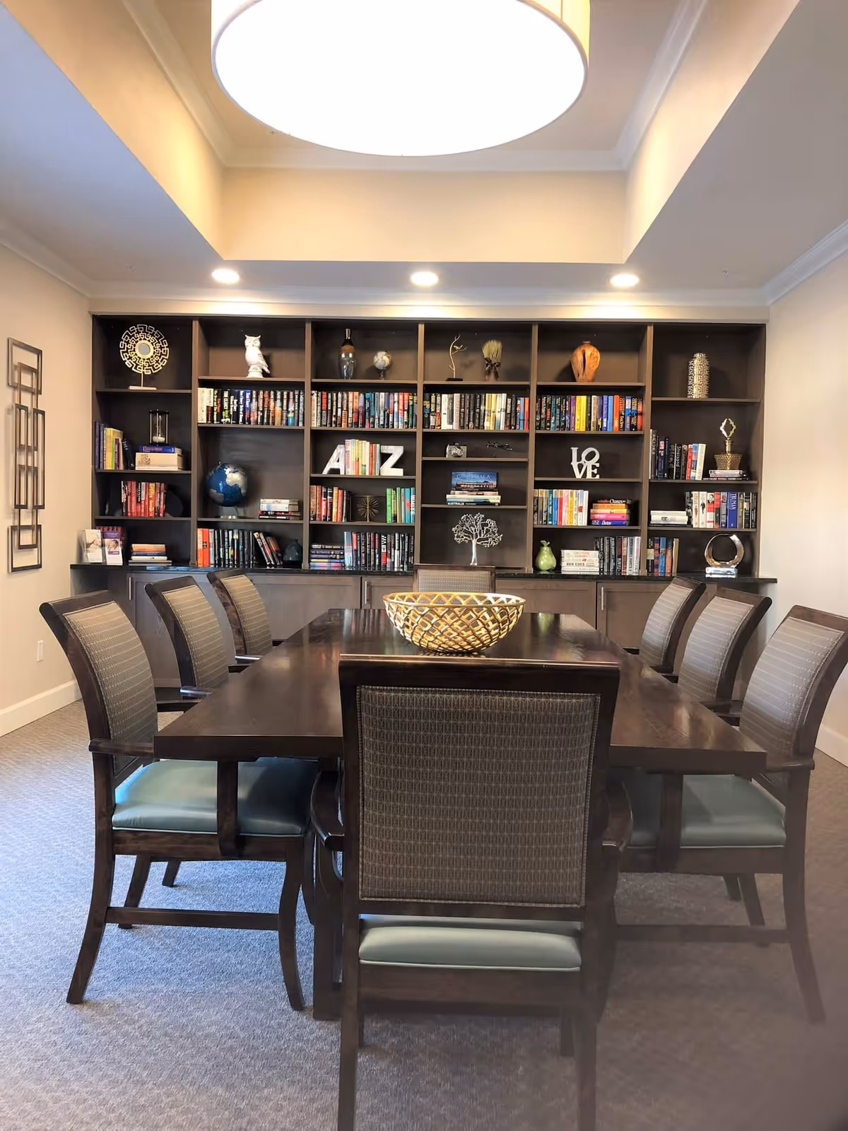 A well-lit room with a large dark wooden table surrounded by eight chairs with cushioned seats and backs. Behind the table is a large built-in bookshelf filled with books and decorative items, including globes, sculptures, and letters spelling 'A to Z' and 'LOVE'. The ceiling has recessed lighting and a large circular light fixture.