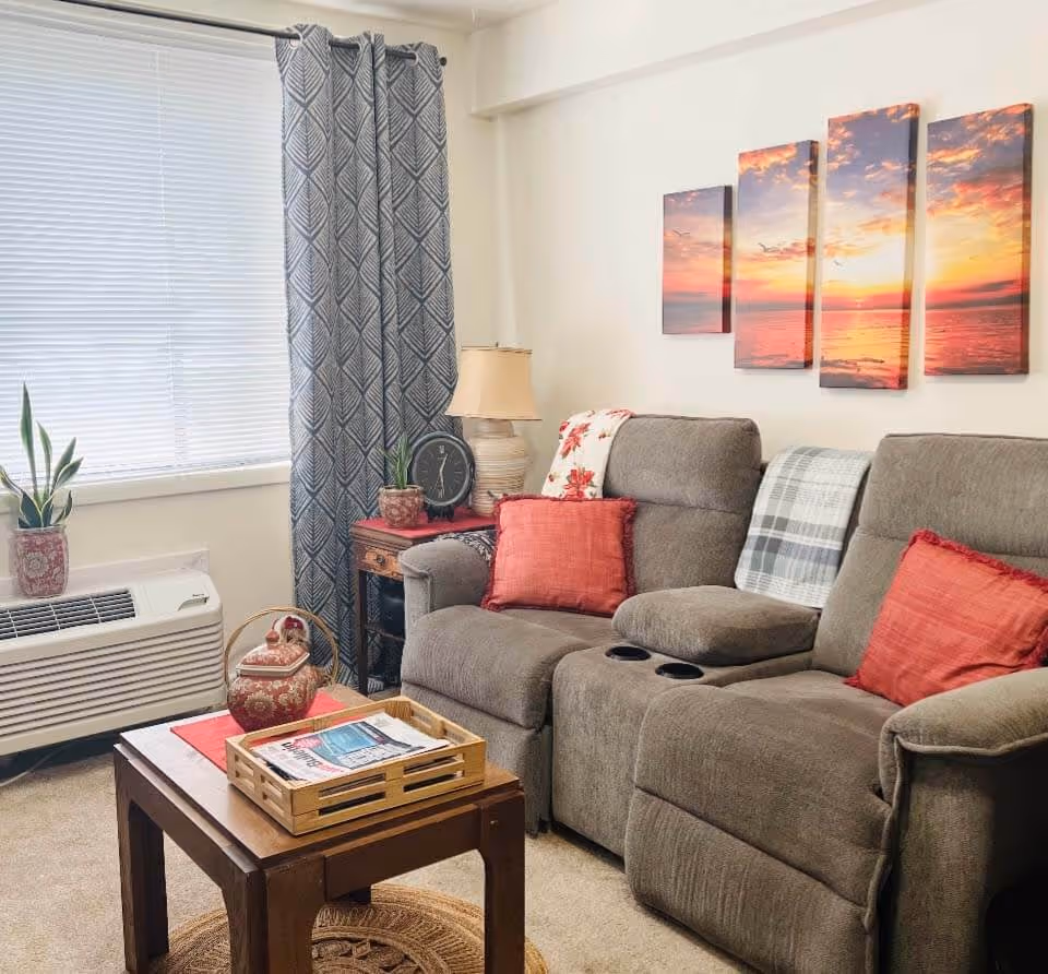 Cozy living room with a gray reclining sofa adorned with red and plaid pillows and blankets. A wooden coffee table holds a decorative tray with magazines and a ceramic teapot. Next to the sofa is a small wooden side table with a lamp, a clock, and a potted plant. A window with blinds and patterned gray curtains lets in natural light. On the wall above the sofa, there are four panels of artwork depicting a sunset over the ocean.