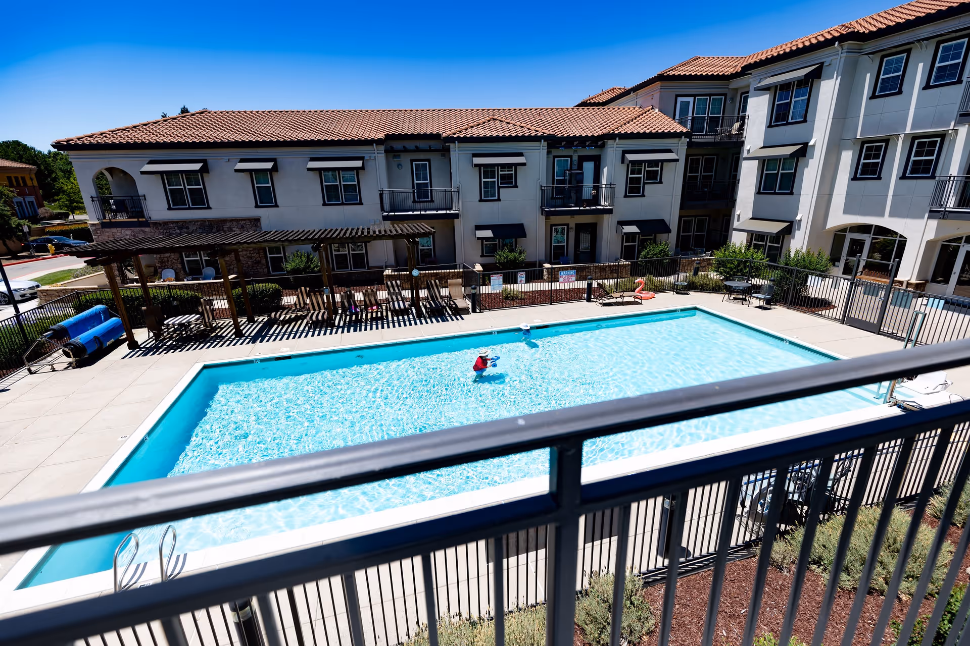 Outdoor swimming pool surrounded by a concrete deck and black metal fence, with a multi-story residential building in the background under a clear blue sky. There are lounge chairs and a pergola on one side of the pool.
