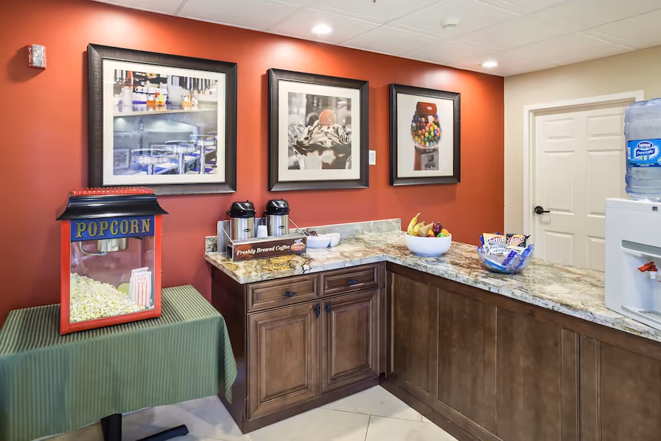 Countertop snack station with a popcorn machine, coffee urns, bowls of fruit and snacks, and a water cooler against a red wall with framed pictures.